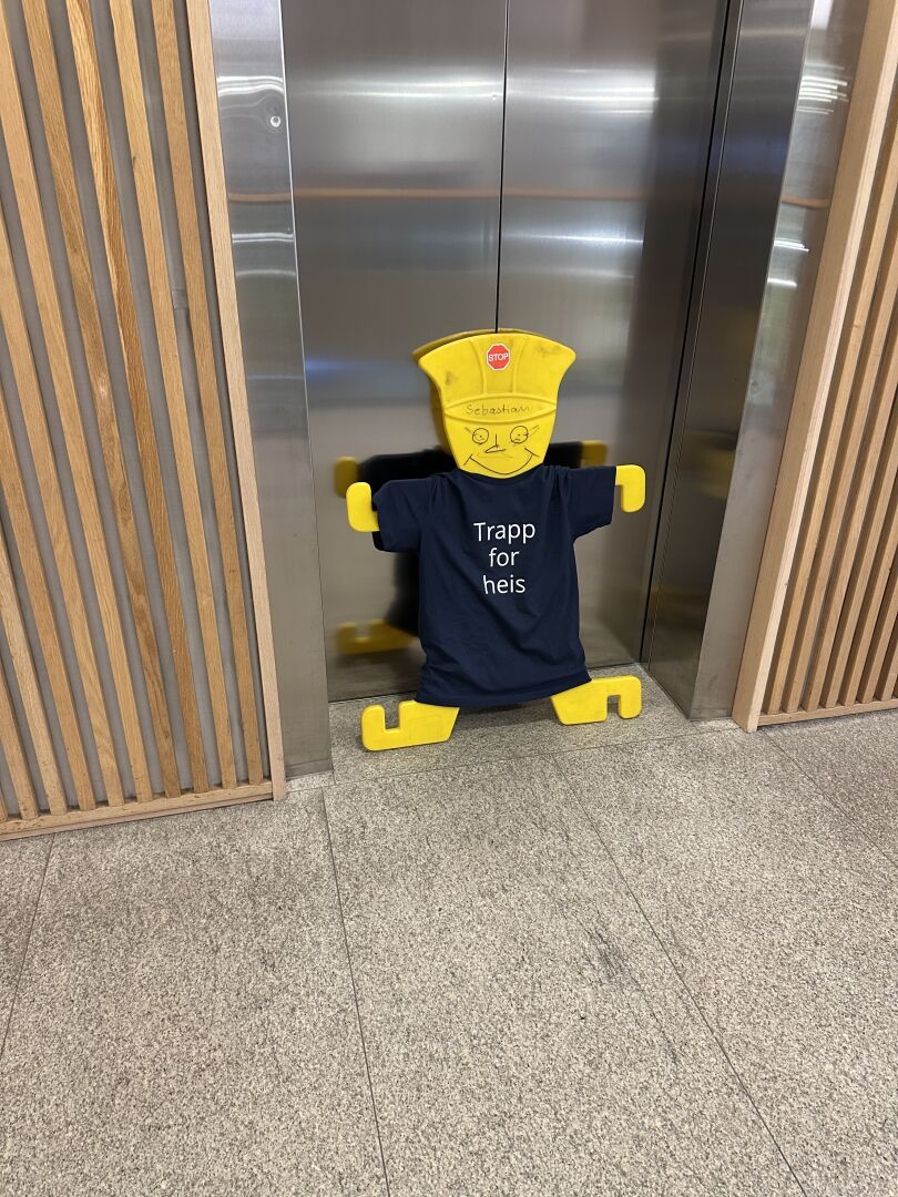 En plastfigur med T-skjorte som har påskriften «trapp for heis» satt foran en heis som er ute av drift.