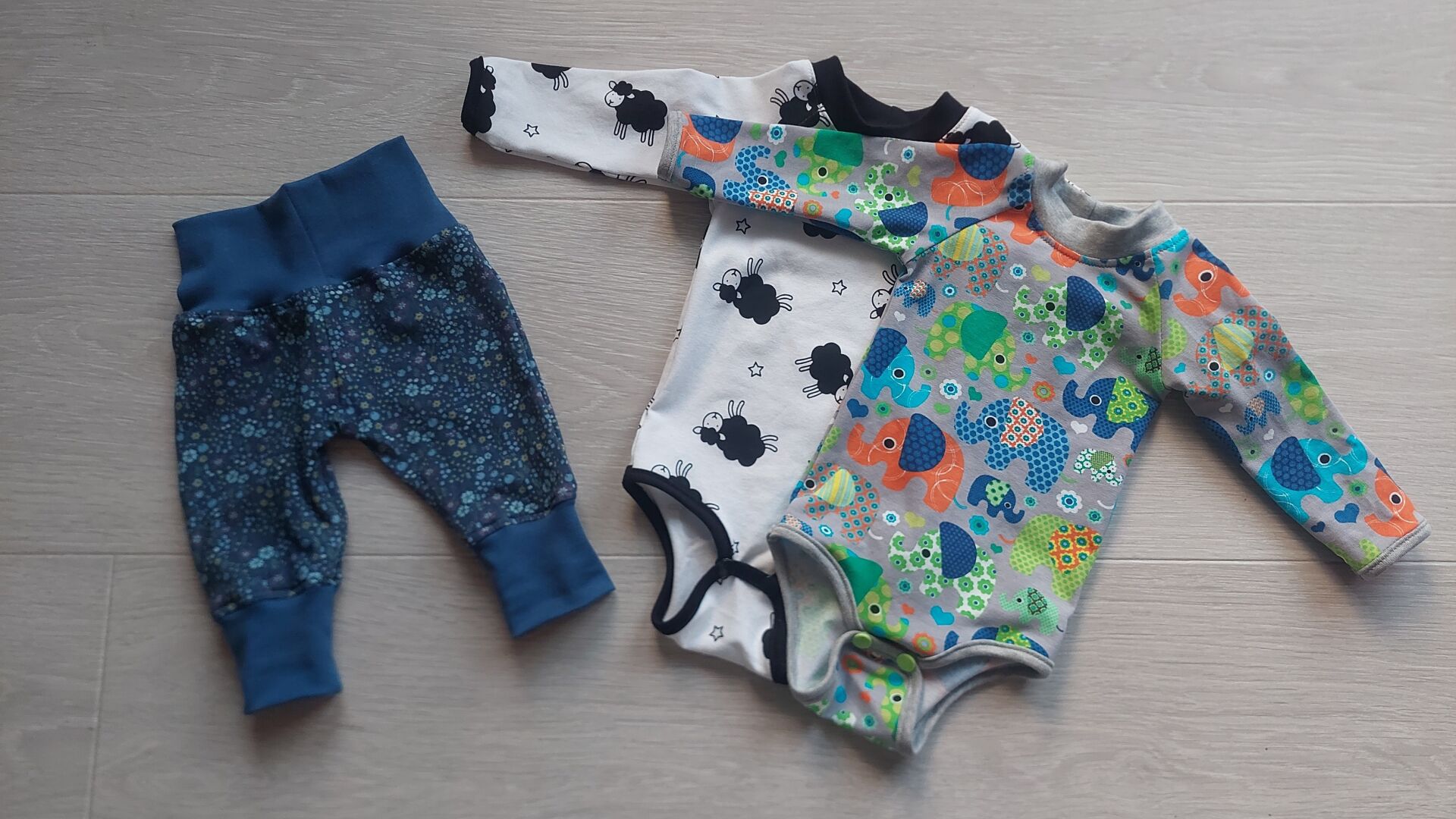 A pair of blue baby pants and two one-pieces lying on the floor. 
The pants have tiny light blue, yellow and red flowers on darker blue. The left one-piece is lying halfway under the right one, and has black sheep on white background, with black trim. The right one-piece has red, green and blue elephants on grey background, with grey trim.