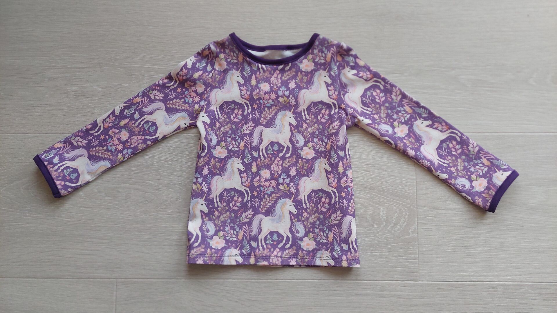 A light purple sweater with white unicorns, and purple and pink flowers, lying on the floor. The trim around the neck and sleeves is dark purple.
