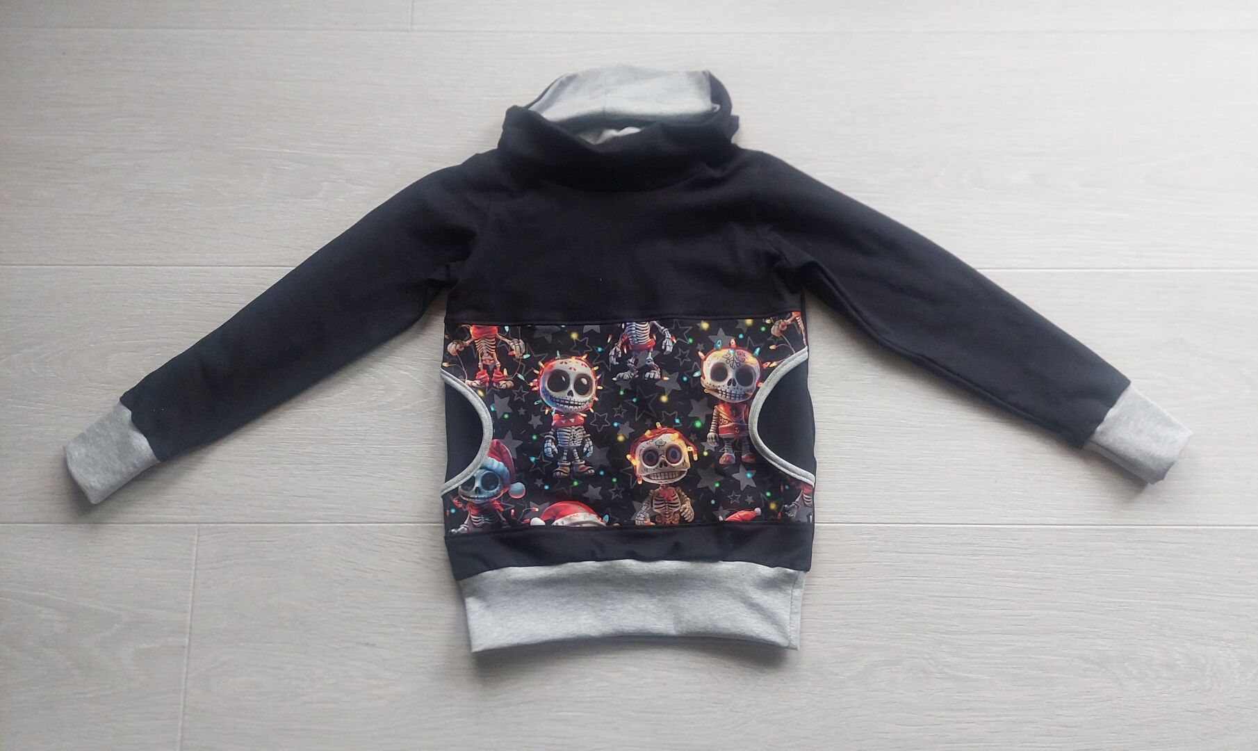 Svart barnegenser med komme på heile framsida og høg krage. Mansjettar, ribb på bolen og fôret på kragen er lys grå. Stoffet på lomma er svart med skjelett og julelys.