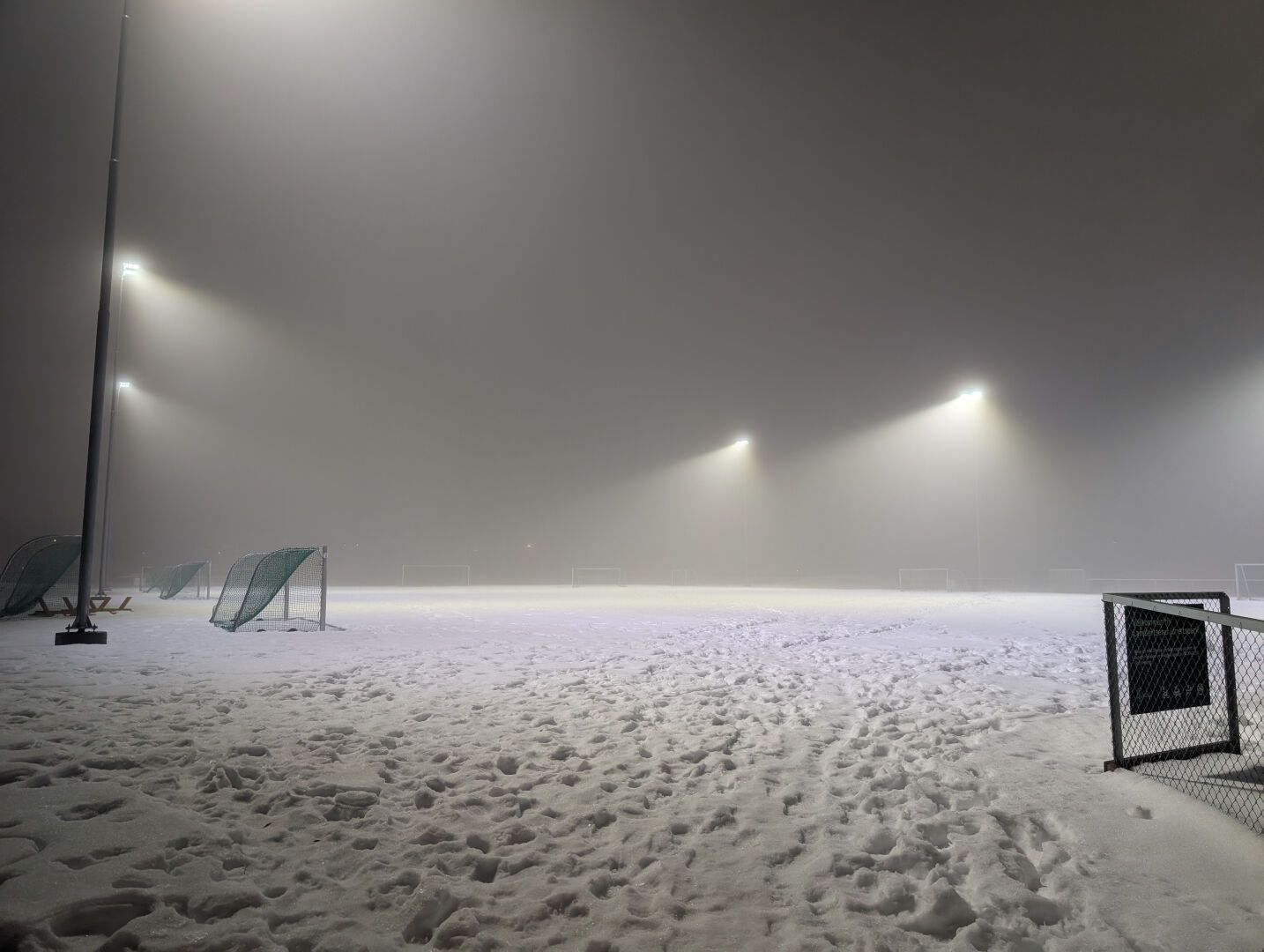 En snøhvit idrettsbane om kvelden, folketom, men opplyst med lyskastere gjennom tåka