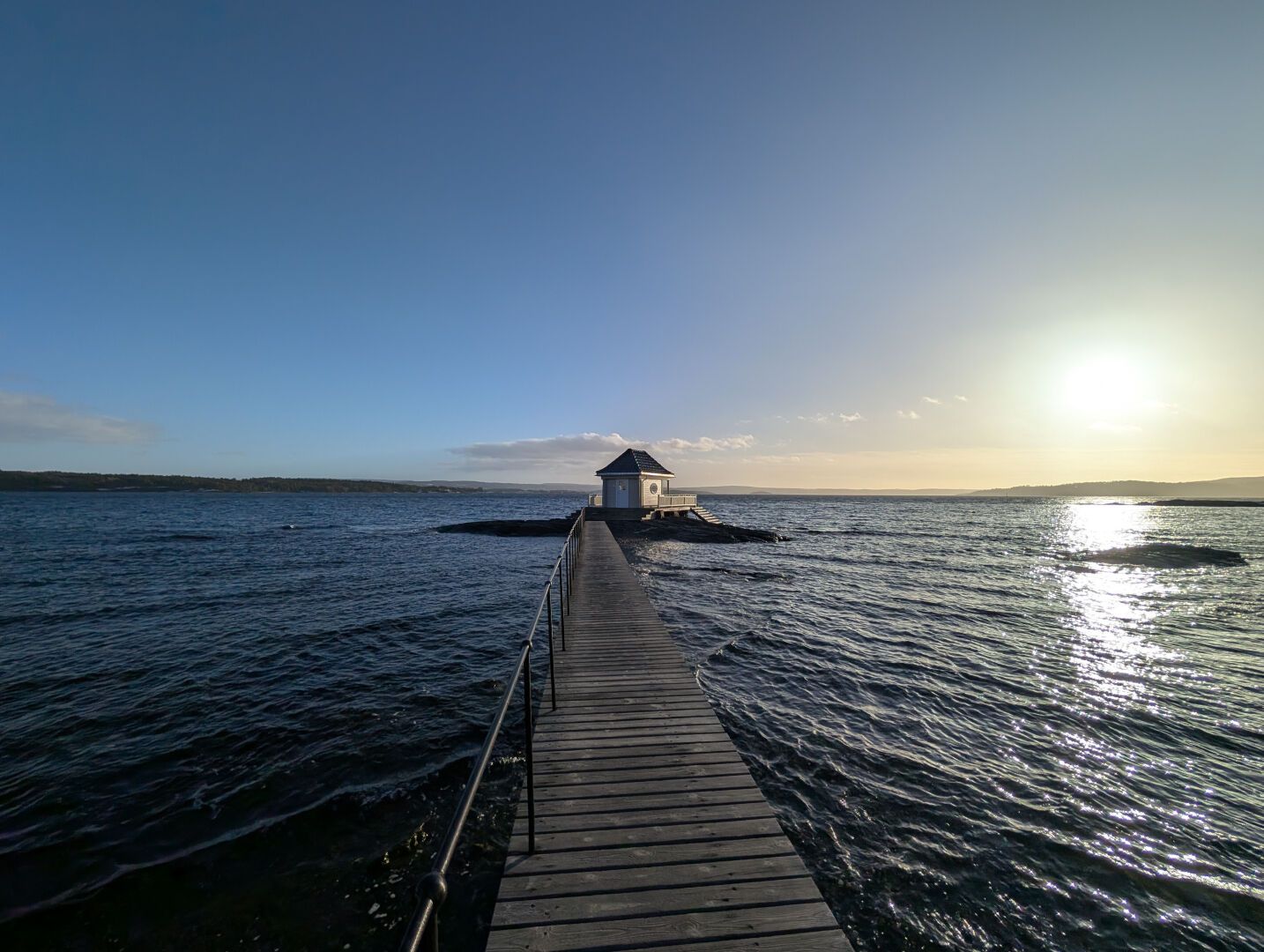sjø og en lang brygge forover som ender i et badehus. Sol og blå himmel